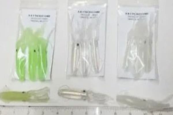 Three packets, each containing fishing lures, positioned above a ruler. The lures in the packets are in various shades of green and white, while five loose lures are placed below the packets in a row.