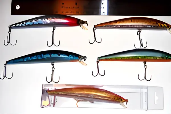 Five fishing lures with treble hooks are displayed on a wall, four arranged above and one in a plastic case below.